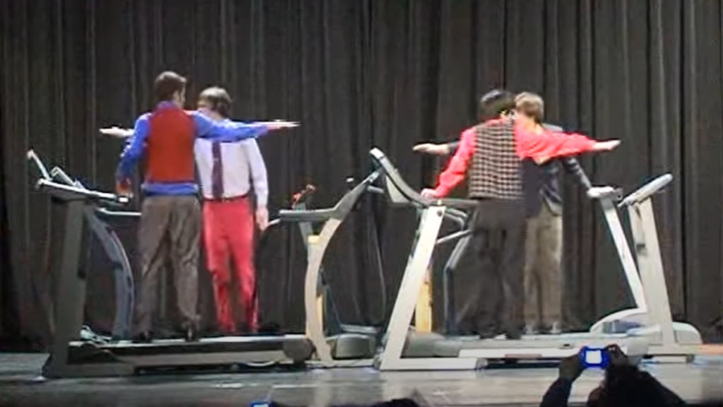 Four treadmills are set up on a stage. Four teens stand on one each. The two closes to the camera have their backs facing us. The two teens behind them face us. They each have their arms stretched out in a cool formation