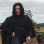 Close up of a man dressed like Professor Snape from Harry Potter. Whatever he is sitting on is partially covered. He is outside