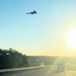 View of a stealth plane in the sky, starting to land. The sky is bright blue, and the sun shines brightly off on the right side of the image