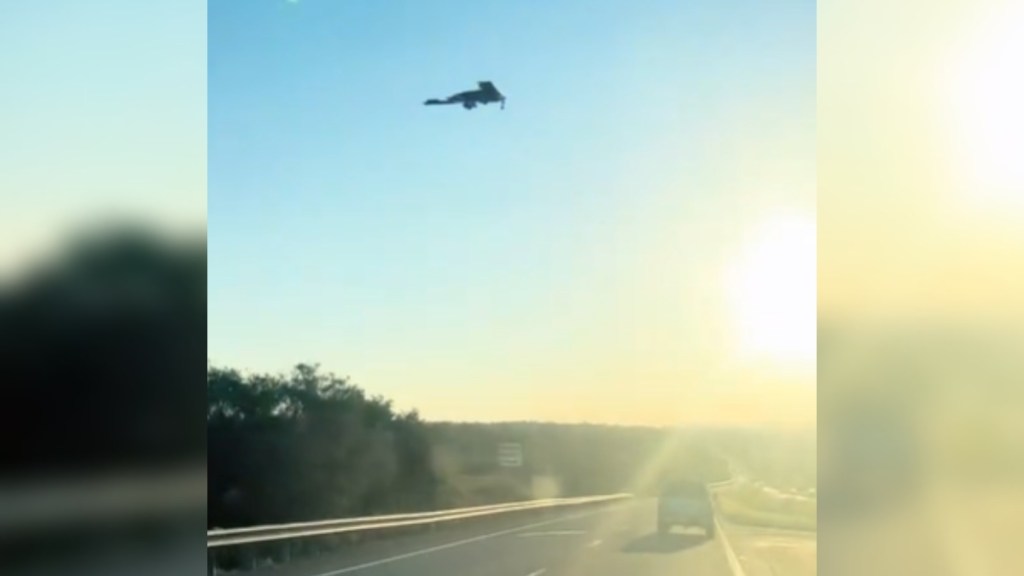 View of a stealth plane in the sky, starting to land. The sky is bright blue, and the sun shines brightly off on the right side of the image