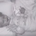 Top-down view of a little boy laying in a crib. The image is in black and white. The little boy smiles, pacifier in his mouth, as he sings. He holds his hands out as if dancing