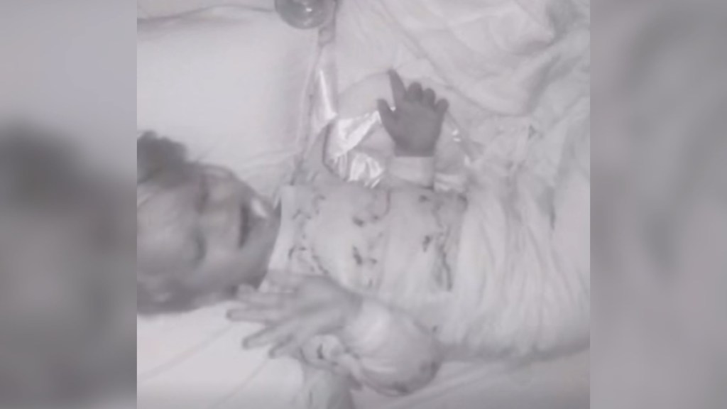 Top-down view of a little boy laying in a crib. The image is in black and white. The little boy smiles, pacifier in his mouth, as he sings. He holds his hands out as if dancing