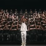 A man in white stands as he orchestrates a large group of people behind him. This group sits in wheelchairs, wearing all black, as they pull off synchronized hand movements