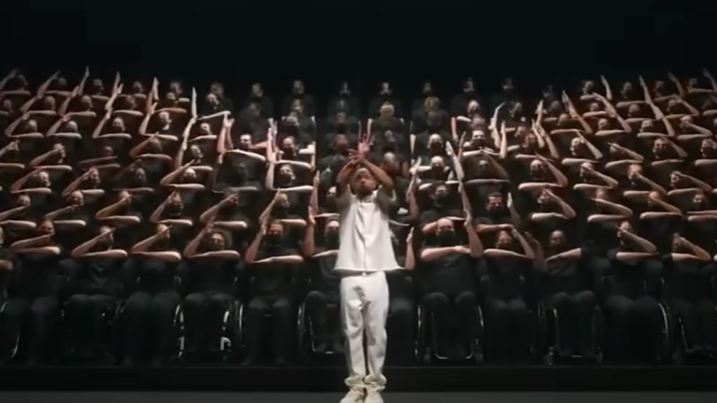 A man in white stands as he orchestrates a large group of people behind him. This group sits in wheelchairs, wearing all black, as they pull off synchronized hand movements