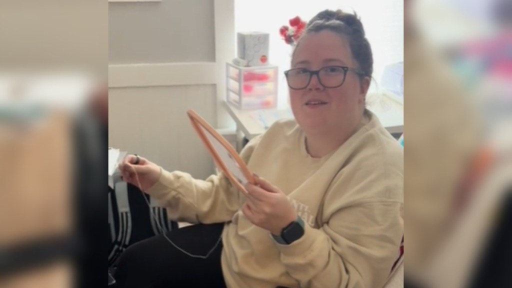 A woman sits in a chair, sewing. She's looking up at the person holding the camera, a look of confusion and curiosity on her face