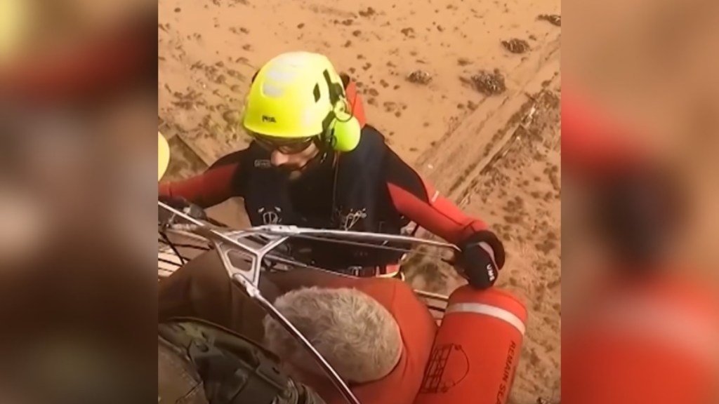 View from above a man hanging off the side of a helicopter. He's assisting as an elderly person is rescued. The elderly person is sat in a large container that's being lifted onto the helicopter. In the distance on the ground, flooding can be seen