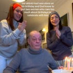 A man sits at a table as two women stand on either side of him. He looks down at the small cake in front of him that has lit birthday candles on them. The two women smile as they clap and sing. Text on the image reads: our airbnb host was alone on his birthday and God new to put two women who care too much about birthdays to celebrate with him