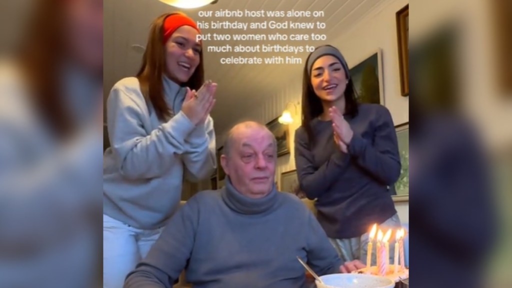 A man sits at a table as two women stand on either side of him. He looks down at the small cake in front of him that has lit birthday candles on them. The two women smile as they clap and sing. Text on the image reads: our airbnb host was alone on his birthday and God new to put two women who care too much about birthdays to celebrate with him