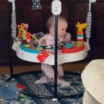 A baby sits in an exersaucer, her legs crossed as she twists in her seat.