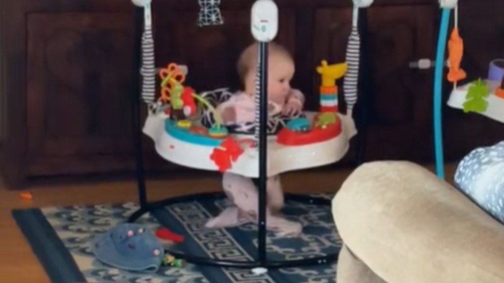 A baby sits in an exersaucer, her legs crossed as she twists in her seat.