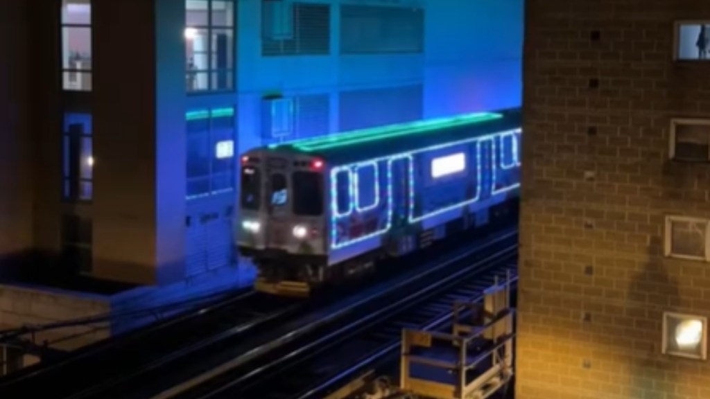 View from an apartment window. At a distance, a train covered in colorful Christmas lights starts to pass by.