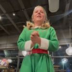 A girl dressed up as a Christmas elf standing up to speak in the middle of a restaurant.