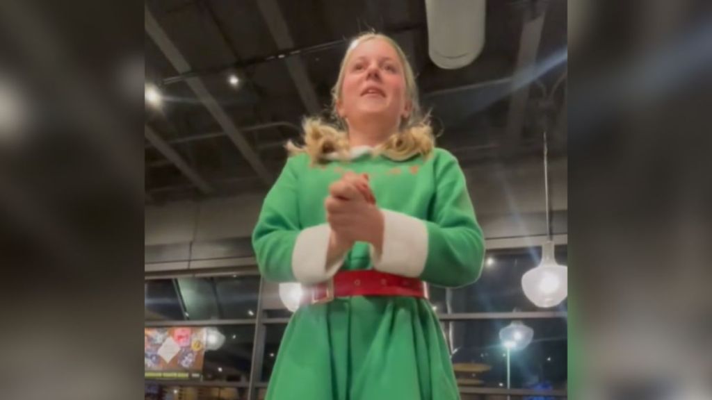 A girl dressed up as a Christmas elf standing up to speak in the middle of a restaurant.