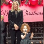 A little girl stands on a stage, mic in hand, as she talks. With one hand she holds a mic and with the other, she holds up one finger. A woman stands next to her, mouth wide open and eyes closed, shocked by what is being said. Behind them on stage is a Christmas banner