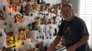 A man looks back at the camera as he adds to his elaborate Christmas village. There are layers to the village with numerous mini buildings placed on fake, fluffy snow