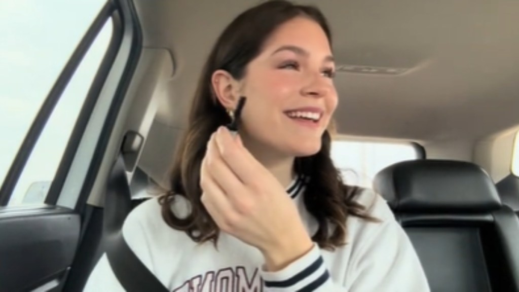 A woman sits in a car, holding mascara in one hand. She wide, a joyful look in her eyes, as she looks over at someone that can't be seen in the photo