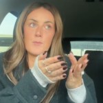 A woman sits in her car, talking to the camera. She has both hands raised and is holding them out forward as she speaks