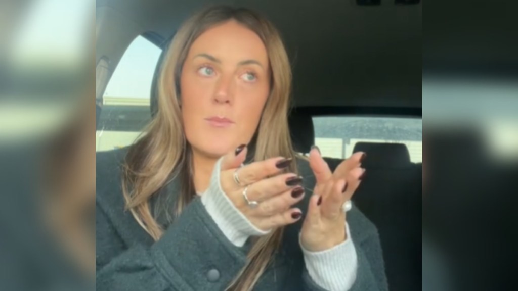 A woman sits in her car, talking to the camera. She has both hands raised and is holding them out forward as she speaks