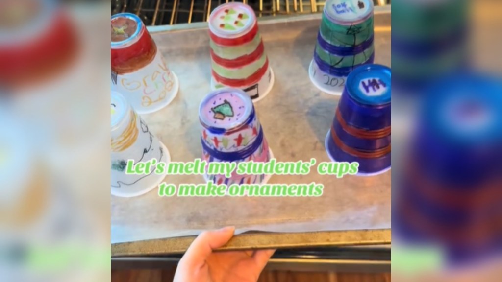 View of a pan being placed into an oven, from the perspective of the person placing it there. On the pan are six plastic cups that are placed upside down and decorated in colorful markers