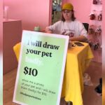 A woman sitting at a table behind a sign that reads, "I will draw your pet badly."