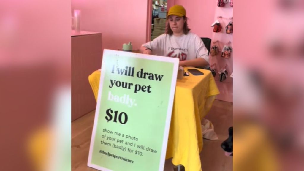 A woman sitting at a table behind a sign that reads, "I will draw your pet badly."