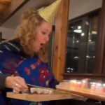 A woman looks down, eyes wide, and mouth open, at the tray she's carrying. On the tray is a birthday cake, with lit candles, that's slipping off