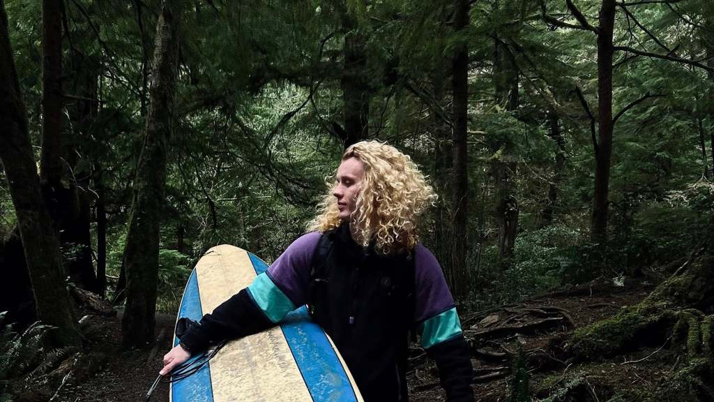 Vlad Siryk, aka Big Water Guy, stands in a forest. He looks to the side as he holds a surf board under one arm.