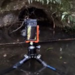 Close up of a camera placed on a tripod. The tripod is in shallow water, facing an opening in a cave