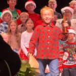 Kids stand on a stage, performing. One kid steps forward, eyes wide. He's got a hammer in one hand and a paper bag in the other. A teacher approaches while wearing a guitar