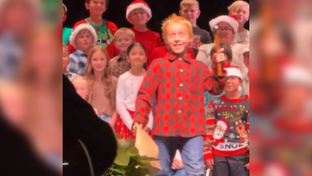 Kids stand on a stage, performing. One kid steps forward, eyes wide. He's got a hammer in one hand and a paper bag in the other. A teacher approaches while wearing a guitar