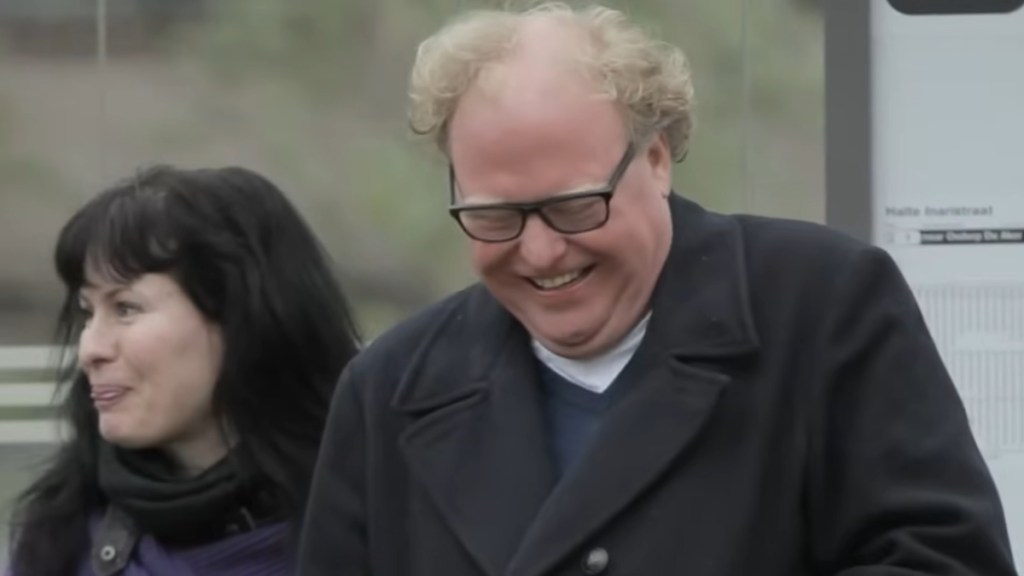 Close up of a man standing outside. He looks down as he smiles and laughs. A woman stands nearby, just a bit behind him. She looks off in the distain, a small smile on her face as she seems to be suppressing a laugh herself.