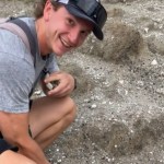 Close up of a man squatting down outside. He looks up at the camera, smiling with a look of surprise on his face.