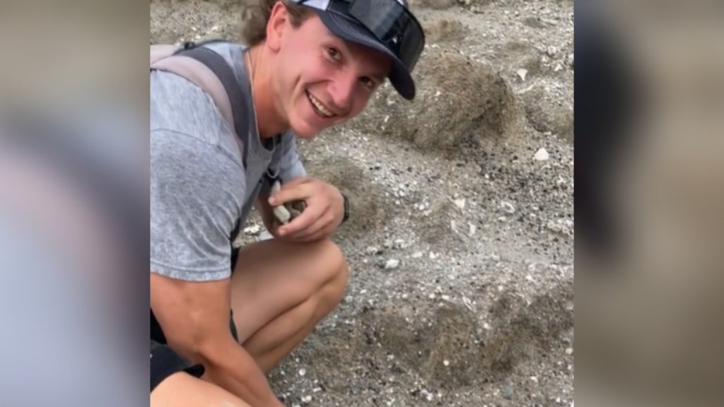 Close up of a man squatting down outside. He looks up at the camera, smiling with a look of surprise on his face.