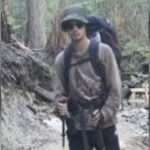 A young man stands in the woods in hiking gear, backpack on his back. He's leaning against walking sticks.