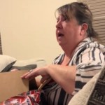 A woman sits on a couch, opening a gift that can't be seen in this image. The woman looks up at someone off to the side that can't be seen. The woman's eyes are wide and mouth is open, shocked