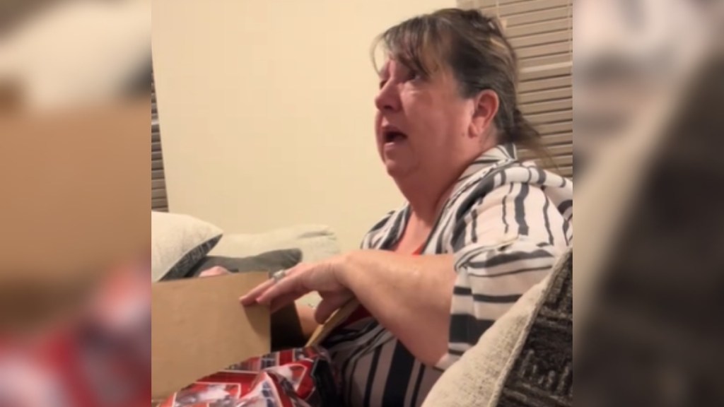 A woman sits on a couch, opening a gift that can't be seen in this image. The woman looks up at someone off to the side that can't be seen. The woman's eyes are wide and mouth is open, shocked