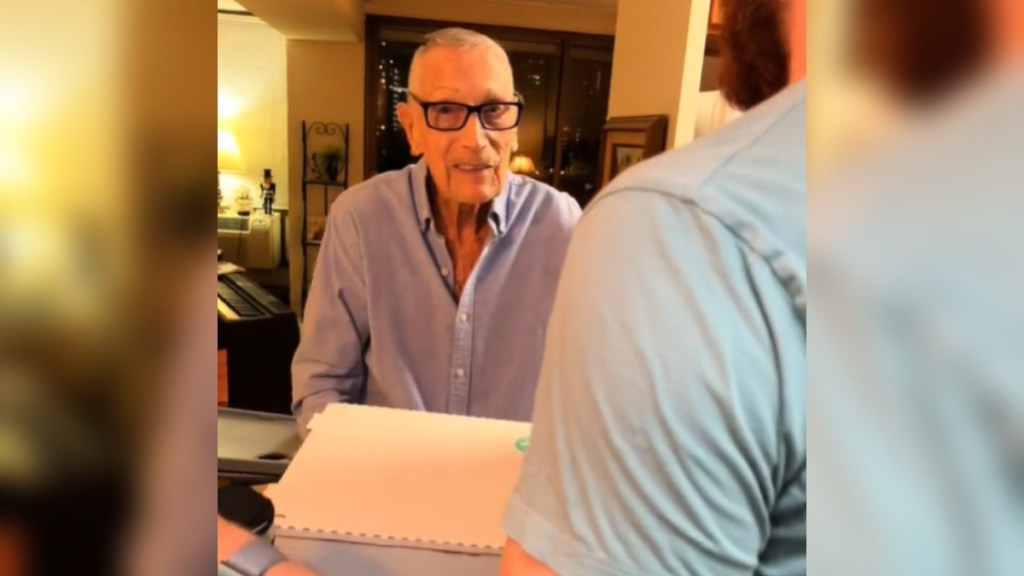 View from behind of a man entering an apartment with a pizza box in hand. He's starting to greet the 98-year-old man in front of him who is smiling at the person with the camera