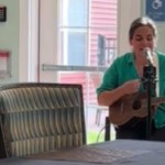 A woman sits as she plays a guitar and sings into a mic. An elderly woman sitting in a wheelchair nearby watches