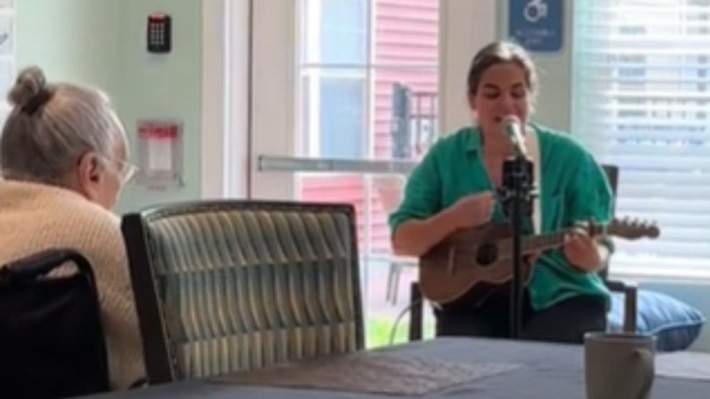 A woman sits as she plays a guitar and sings into a mic. An elderly woman sitting in a wheelchair nearby watches