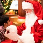 A little kid looks at Santa Claus wide-eyes. Santa sits in a chair, looking back. He holds the kid's letter in his hands.