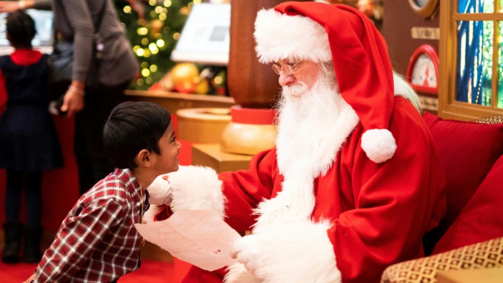 A little kid looks at Santa Claus wide-eyes. Santa sits in a chair, looking back. He holds the kid's letter in his hands.