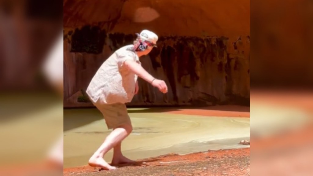 View from behind of a man in a cave at a canyon in Utah. He's moving an arm back to skip a rock on the water near him