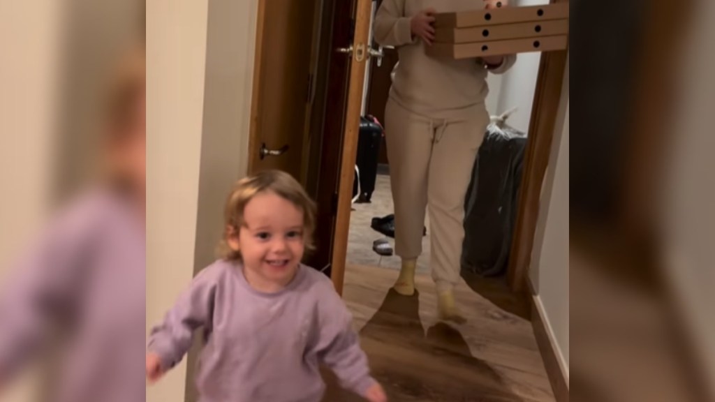 A toddler excitedly runs toward the person capturing this image. Just behind the toddler is someone holding boxes of pizza