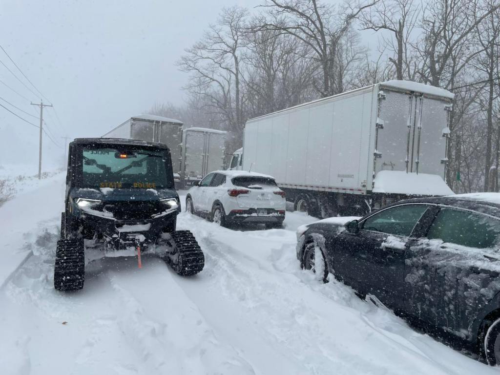 A state trooper vehicle parked in the snow next to stranded cars and trucks. 