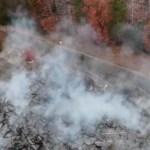 Aerial view of a parking lot with a fire beneath. Smoke rises from the parking lot. Nearby are colorful fall trees