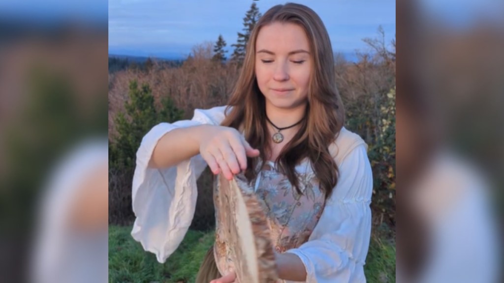 Close up of a woman standing outside. She looks down as she starts to turn a piece of wood around. Mid-movement, a glimpse of the woodburning art she created can be seen, but we can't see much yet