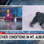 Image shows Bob Herzog making snow angels during a live weather broadcast as inside anchors look on.