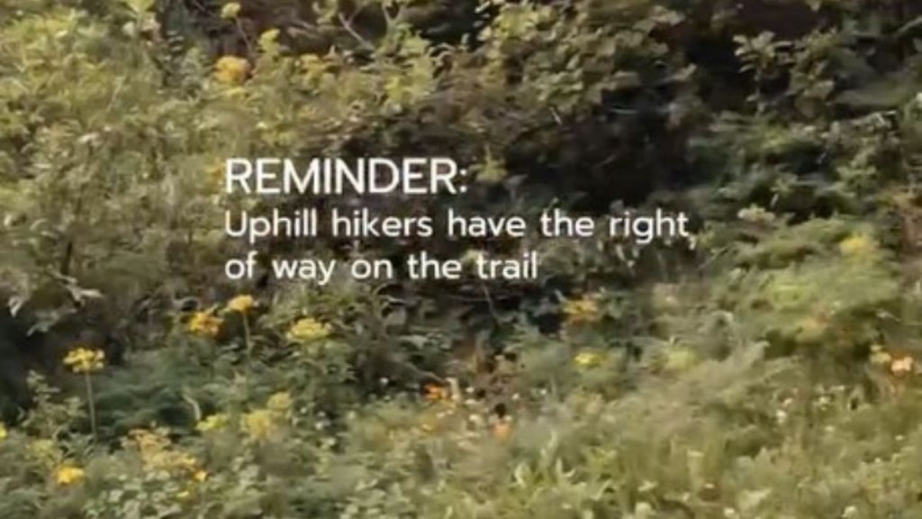 Image shows mountain foliage with a hiking rule, "REMINDER: Uphill hikers have the right of way on the trail."