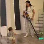 A mom stands with a vacuum in hand. She looks down in shock and awe at her baby who is starting to stand