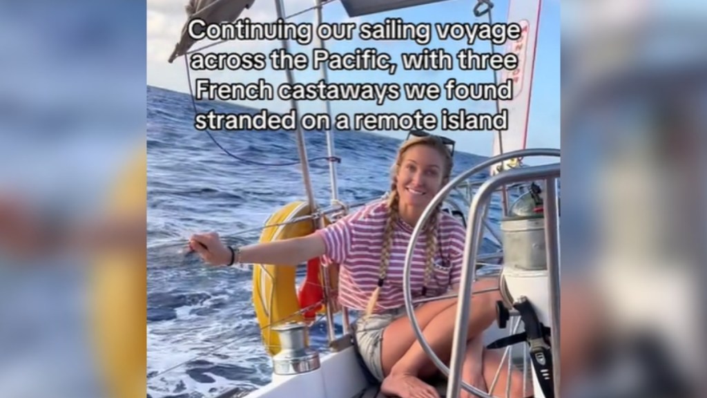 A woman sits on a sailboat. She smiles at the camera with a look of uncertainty in her eyes. Text on the image reads: Continuing our sailing voyage across the Pacific, with three French castaways we found stranded on a remote island.